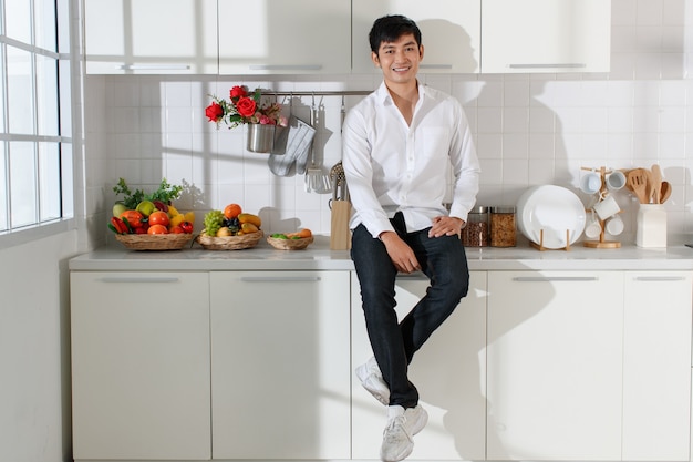 Młody atrakcyjny Azjata ubrany w białą koszulę i dżinsy siedzący w białej kuchni