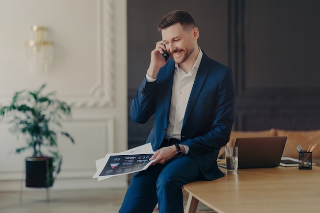 Młody atrakcyjny biznesmen w formalnym ciemnoniebieskim garniturze rozmawia przez telefon w biurze, uśmiechając się, słysząc o dobrych wynikach swojego zespołu, siedząc na biurku z laptopem, szklanką wody i ołówkami