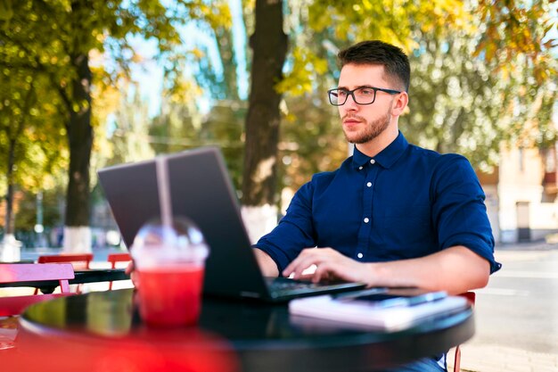 Młody atrakcyjny freelancer hipster pracujący zdalnie na laptopie w kawiarni na świeżym powietrzu biznesmen przeglądający