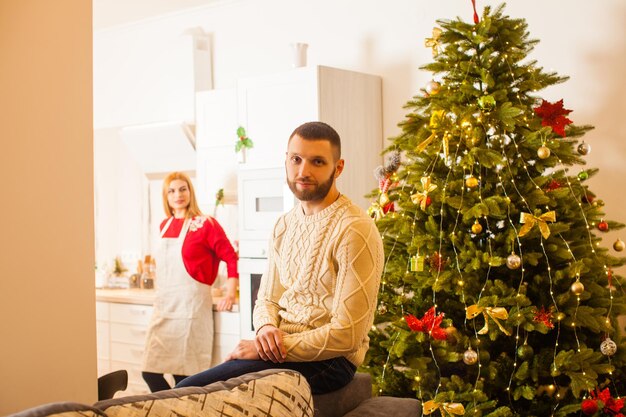 Młody atrakcyjny nieogolony mężczyzna w sweter z dzianiny, siedzący na tylnej kanapie w swoim domu. Udekorowana choinka blisko niego, młoda żona na tle, blured.