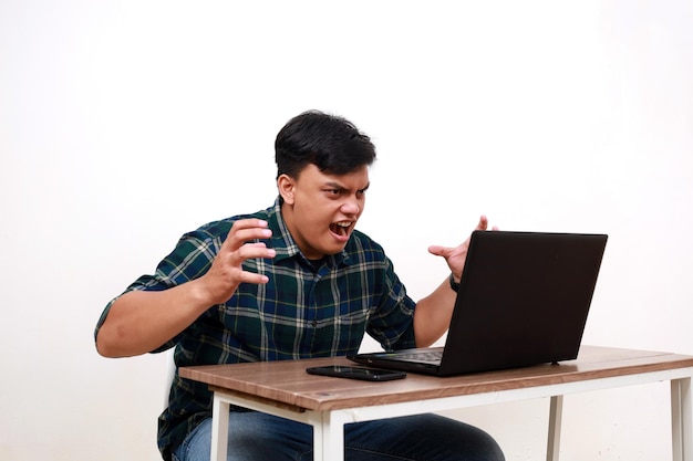 Młody Azjat używający laptopa z gniewnym wyrazem twarzy i gestem