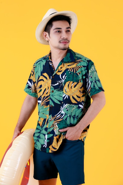 Młody Azjata w kolorowej hawajskiej koszuli z nadrukiem uśmiecha się ze szczęścia, trzymając kółko do pływania. Portret studio pół ciała na żółtym tle. Koncepcja podróży wakacje letnie.