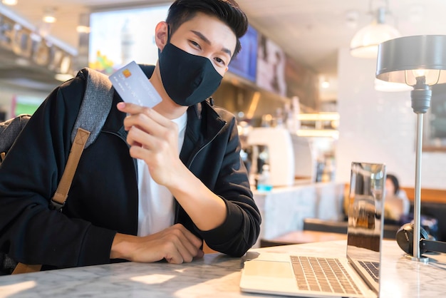 Młody azjatycki atrakcyjny męski podróżnik noszący maskę na twarz chroniący przed wirusami trzymaj kartę kredytową na zakupy online z laptopem w kawiarni restauracja pomysły biznesowe nowy normalny styl życia