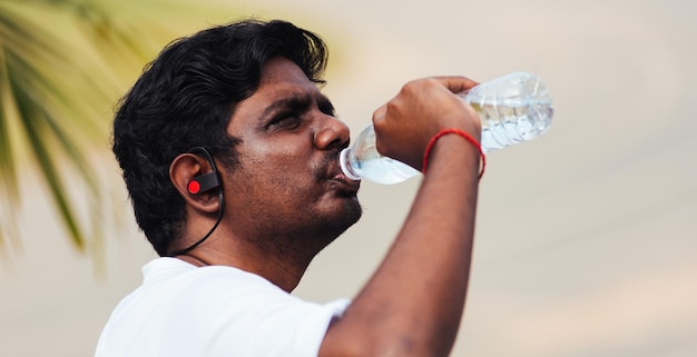 Młody azjatycki biegacz sportowy z bliska, czarny mężczyzna, nosi słuchawki sportowca, który pije wodę z butelki po bieganiu w parku zdrowia na świeżym powietrzu, koncepcja treningu zdrowych ćwiczeń