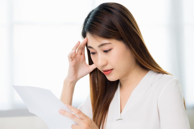Młody azjatycki biznes kobieta ręką dotykając głowy patrząc na papierkową robotę lub rachunki, ona czuje się przygnębiony stres ból głowy być zmęczony od długiej pracy z laptopem, ona objaw zespołu biurowego.