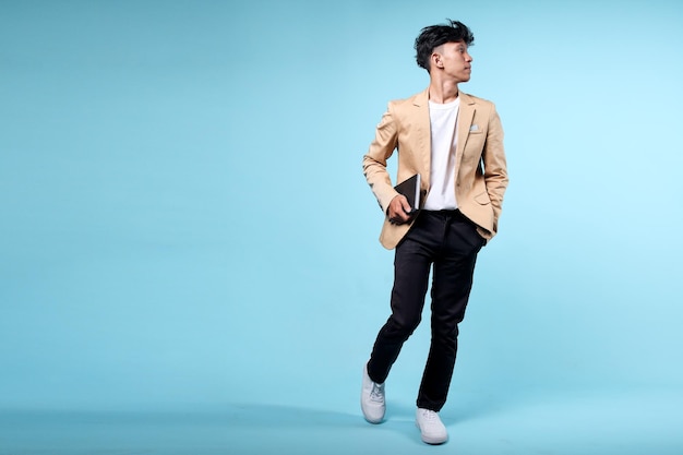 Młody azjatycki biznesmen w stylowych ubraniach w pozycji chodzenia i patrząc na boki na niebieskim tle