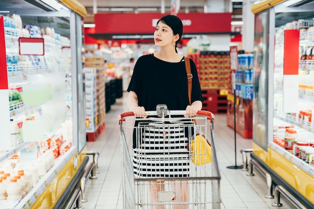 Młody azjatycki chiński uroczy kobieta w ciąży pchanie koszyka z półkami produktów spożywczych na boku. piękne przyszłe macierzyństwo kupując jedzenie z wózkiem w sklepie. elegancki rodzic w supermarkecie