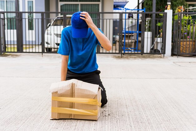 Młody azjatycki dostawca w niebieskim mundurze emocjonalnie spadający kurier trzyma uszkodzony karton przed drzwiami domu, wypadek złej przesyłki transportowej lub koncepcja usługi dostawy złej jakości