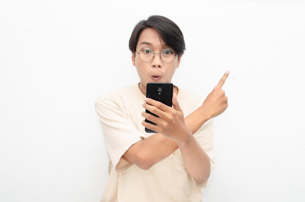 młody azjatycki mężczyzna w beżowej koszulce trzymający i pokazujący ekran telefonu ze szczęśliwą, euforyczną, zszokowaną twarzą