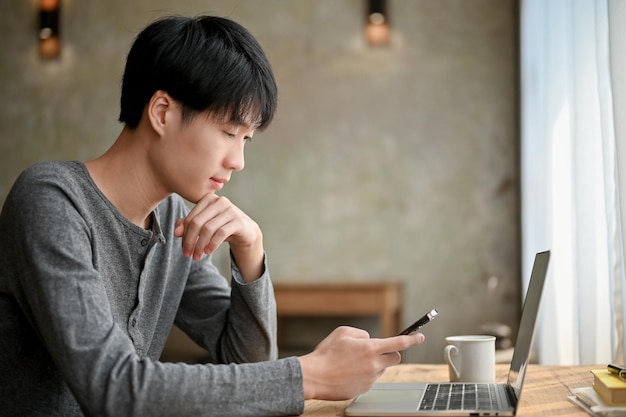 Młody azjatycki mężczyzna w zamyśleniu myśli o czymś, patrząc na ekran swojego smartfona