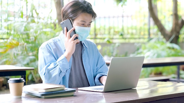 Młody azjatycki student noszący maskę ochronną rozmawiający telefon i pracujący z laptopem