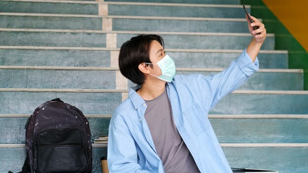 Młody azjatycki student uniwersytetu trzymający telefon komórkowy podczas rozmowy wideo podczas noszenia maski ochronnej