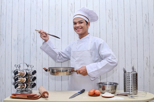 Młody azjatycki szef kuchni w białym toczku i fartuchu z patelnią i drewnianą szpatułką, degustujący jedzenie.