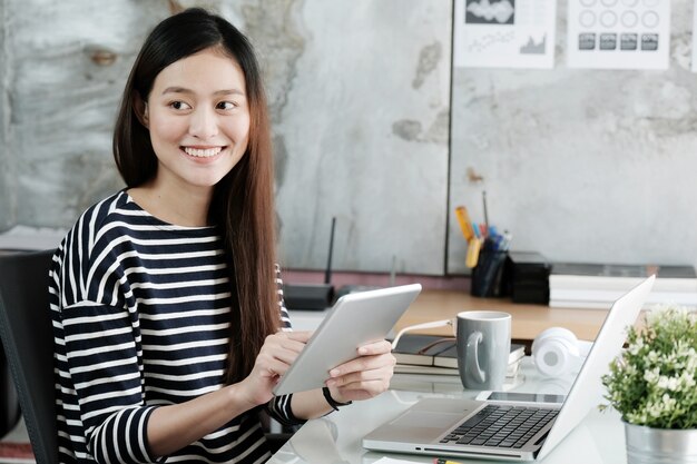 Młody azjatykci bizneswoman pracuje z pastylką i laptopem z uśmiechniętą twarzą