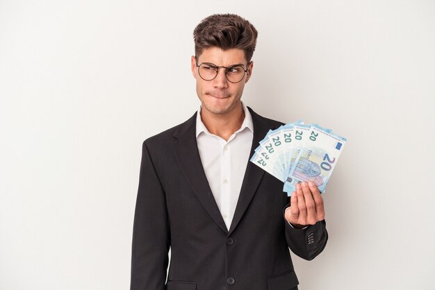 Młody biznes kaukaski mężczyzna trzyma banknoty na białym tle zdezorientowany, czuje się wątpliwy i niepewny.