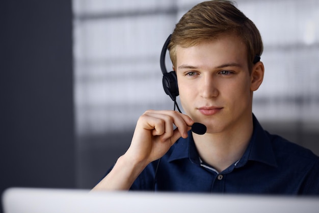 Młody biznesmen blond za pomocą zestawu słuchawkowego i komputera w pracy. Startup oznacza ciężką pracę i brak czasu na osiągnięcie sukcesu.