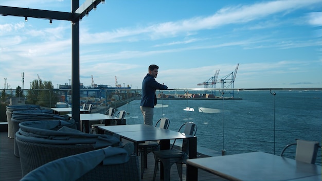 Młody biznesmen odpoczywa na zewnątrz, ciesząc się widokiem na morze na szklanym tarasie kawiarni