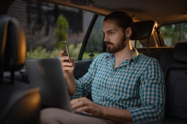 Młody Biznesmen Pewnie Trzyma W Ręku Smartfon I Wysyła Sms-y Na Tylnym Siedzeniu W Samochodzie.