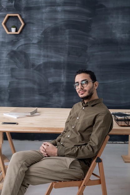 Młody biznesmen poważny w casualwear i okularach siedzi na krześle przy stole na przestrzeni tablicy