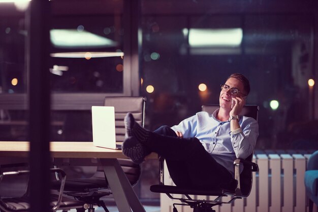 Młody biznesmen przy użyciu telefonu komórkowego podczas pracy na laptopie w nocy w ciemnym biurze.