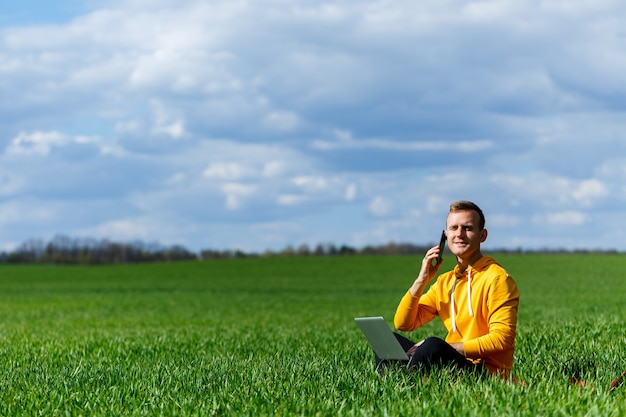 Młody Biznesmen Siedzi Na Zielonej Trawie Rozmawia Przez Telefon I Przy Użyciu Komputera Przenośnego. Przystojny Mężczyzna Pracujący Z Komputerem W Parku W Słoneczny Letni Dzień. Koncepcja Pracy Freelancer