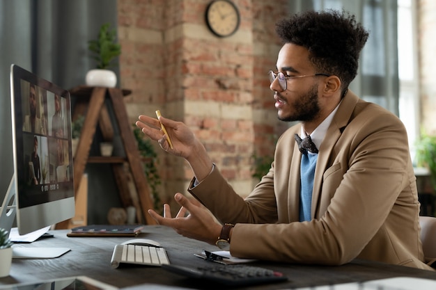 Młody biznesmen siedzi w swoim miejscu pracy przed monitorem komputera i omawia ze swoimi partnerami momenty pracy podczas spotkania online