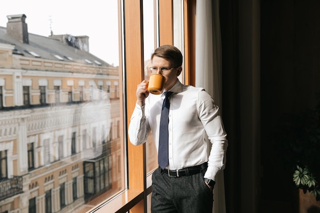 Młody biznesmen stoi przy filiżance kawy przy oknie swojego biura