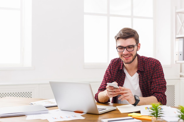 Młody biznesmen szczęśliwy za pomocą telefonu komórkowego przez laptopa w nowoczesnym wnętrzu biurowym biały.