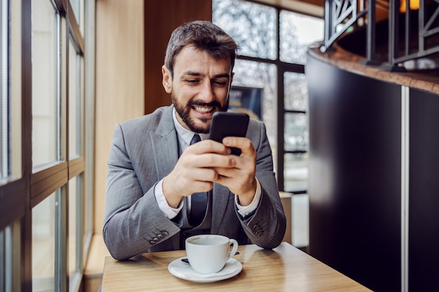 Młody biznesmen uśmiechający się siedzi w kawiarni na przerwę na kawę i wpisując wiadomość na smartfonie.