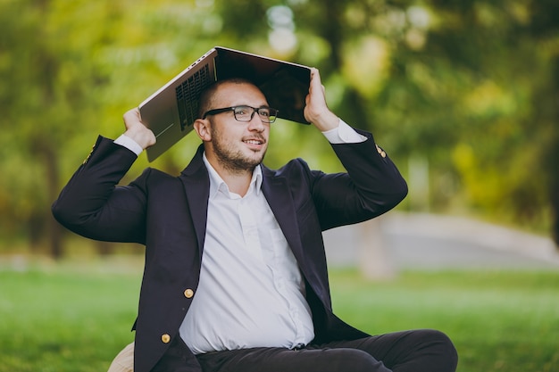 Młody biznesmen w białej koszuli, klasyczny garnitur, okulary. Człowiek siedzieć na miękkiej pufie pod przykryciem laptopa pc komputer w parku miejskim na zielonym trawniku na zewnątrz na przyrodzie. Mobilne biuro, koncepcja biznesowa.