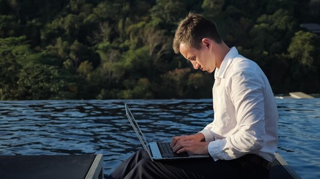 Młody biznesmen w białej koszuli pisze na laptopie przed zielonym leśnym wzgórzem i falującym niebieskim zbliżeniem basenu hotelowego