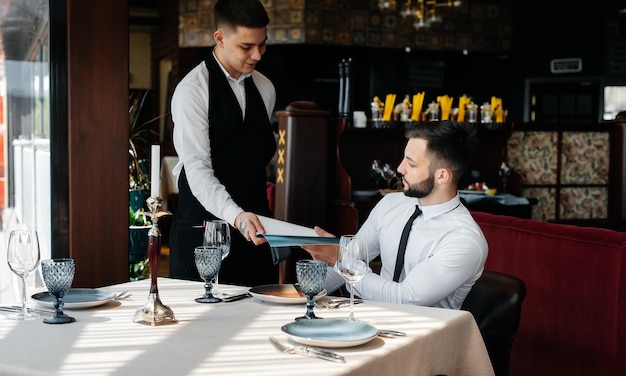 Młody biznesmen w eleganckiej restauracji przegląda menu i składa zamówienie młodemu kelnerowi w stylowym fartuchu Obsługa klienta Obsługa kelnerska w restauracji