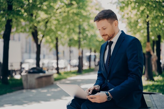 Młody Biznesmen W Niebieskim Garniturze Przegląda Dane I Informacje Na Swoim Laptopie, Surfując Po Internecie, Pracując Zdalnie Nad Projektem, Siedząc Na ławce W Parku Miejskim Z Zielonymi Drzewami W Letni Dzień