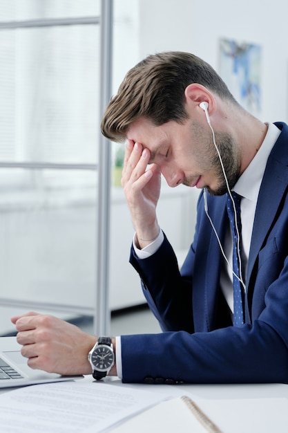 Młody Biznesmen W Słuchawkach Zmęczony Pracą Biurową Siedzi Przy Biurku I Pocieranie Czoła Czując Ból Głowy