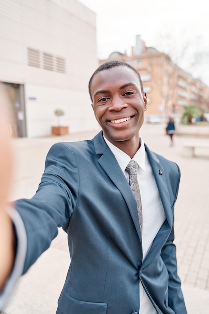 Młody biznesmen z Afryki, ubrany w formalny garnitur, biorąc selfie poza centrum pracy uśmiechnięty