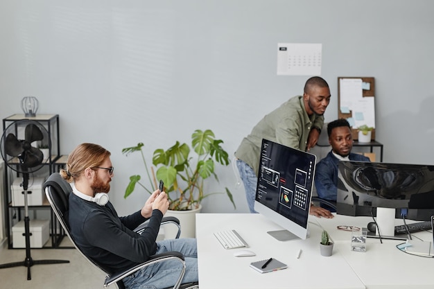 Zdjęcie młody biznesmen ze smartfonem siedzący przy biurku przed ekranem komputera przeciwko dwóm afroamerykańskim współpracownikom