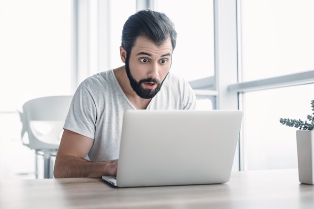 Młody biznesmen zestresowany siedzący z laptopem ze zszokowanym wyrazem twarzy zaskoczony biznesmenem