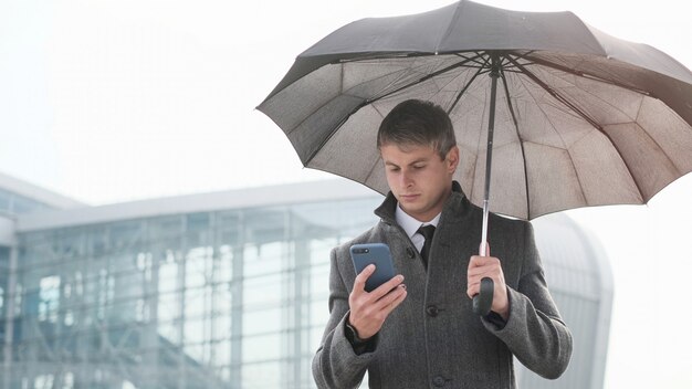 Młody Biznesmena Mienia Parasol I Używać Mądrze Telefon W Deszczowym Dniu.