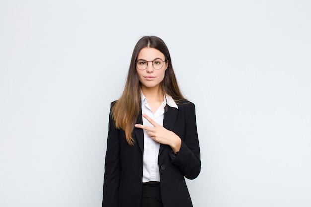 Młody bizneswoman czuje się szczęśliwy, pozytywny i pomyślny, z ręką robi v kształtowi nad klatką piersiową, pokazuje zwycięstwo lub pokój nad biel ścianą