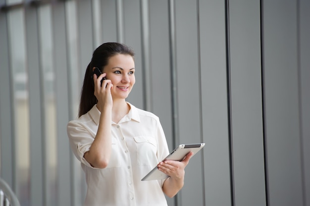 Młody bizneswoman opowiada na telefonie komórkowym podczas gdy stojący okno w biurze. Piękna młoda modelka w biurze.