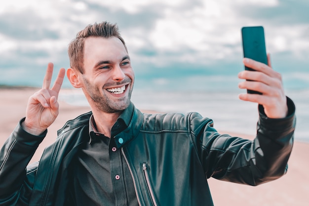 Młody blogger robi selfie lub przesyła strumieniowo wideo na plaży za pomocą czarnego smartfona.