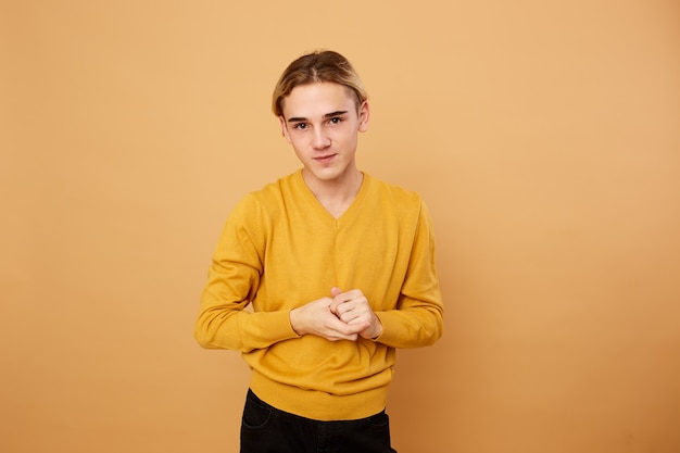 Młody blond facet ubrany w żółty sweter pozuje w studio na beżowym tle.