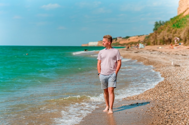 Młody blondyn spaceruje po plaży w pobliżu lazurowego oceanu lub morza Pojęcie szczęśliwego lata