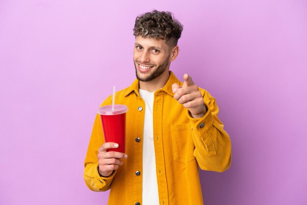 Młody blondyn trzymający napój gazowany na fioletowym tle wskazujący przód ze szczęśliwym wyrazem twarzy
