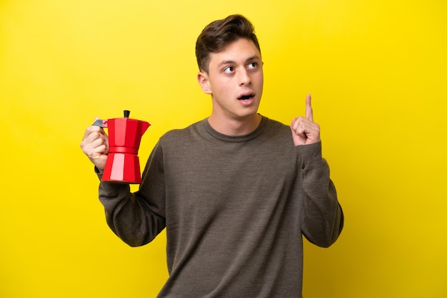 Młody Brazylijczyk trzymający dzbanek do kawy na żółtym tle, myślący o pomyśle wskazującym palec w górę