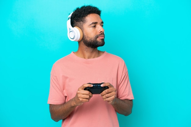 Młody brazylijski mężczyzna bawiący się kontrolerem gier wideo na białym tle na niebieskim tle, patrząc w bok