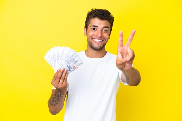 Młody brazylijski mężczyzna biorący dużo pieniędzy na białym tle na żółtym tle uśmiechający się i pokazujący znak zwycięstwa