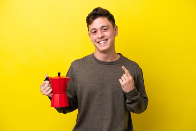 Młody brazylijski mężczyzna trzymający dzbanek do kawy na białym tle na żółtym tle dający gest kciuka w górę