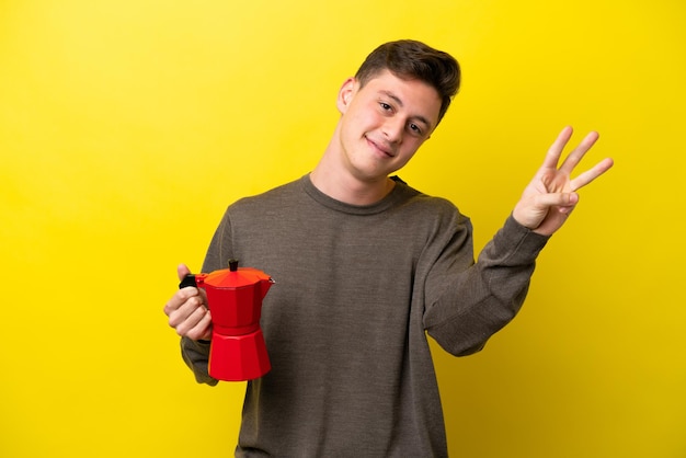 Młody brazylijski mężczyzna trzymający dzbanek do kawy na białym tle na żółtym tle szczęśliwy i liczący trzy palcami
