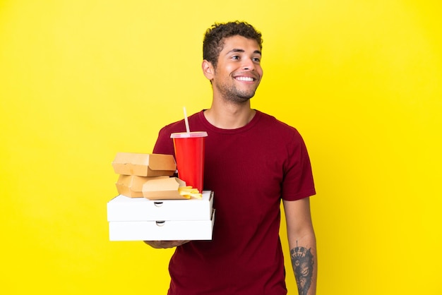 Zdjęcie młody brazylijski mężczyzna trzymający pizze i hamburgery na białym tle, myślący o pomyśle, patrząc w górę